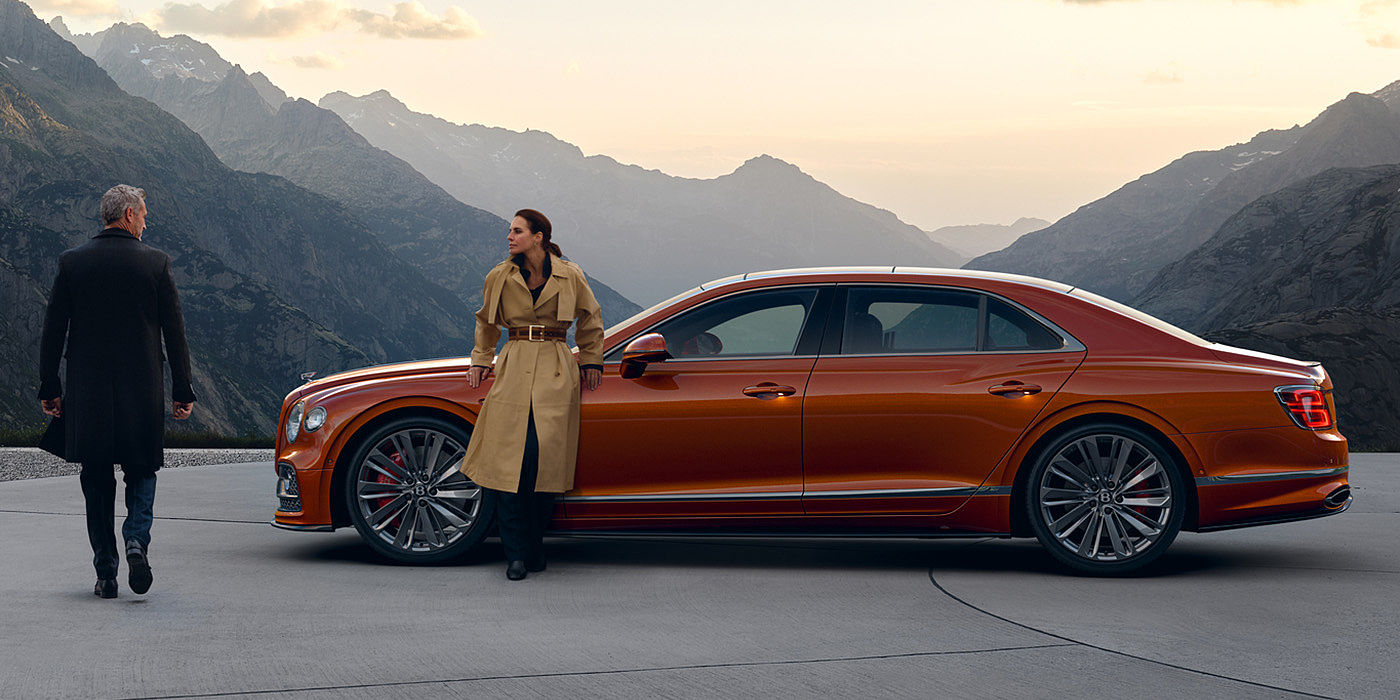 Gohm Sportwagen GmbH | Bentley Singen Bentley Flying Spur Speed parked in Orange Flame coloured exterior parked, with mountainous background and two people in view.