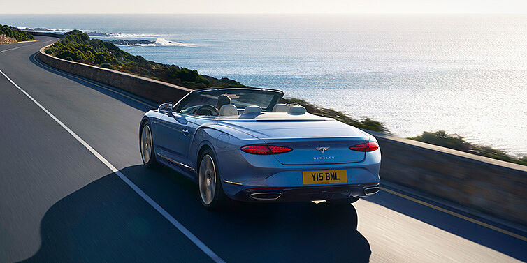 Gohm Sportwagen GmbH | Bentley Singen New Bentley Continental GTC Azure convertible rear view in Light Windsor blue paint with 22 inch ten spoke wheels driving dynamically on an ocean road in the sunshine