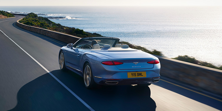 Gohm Sportwagen GmbH | Bentley Singen New Bentley Continental GTC Azure convertible rear view in Light Windsor blue paint with 22 inch ten spoke wheels driving dynamically on an ocean road in the sunshine